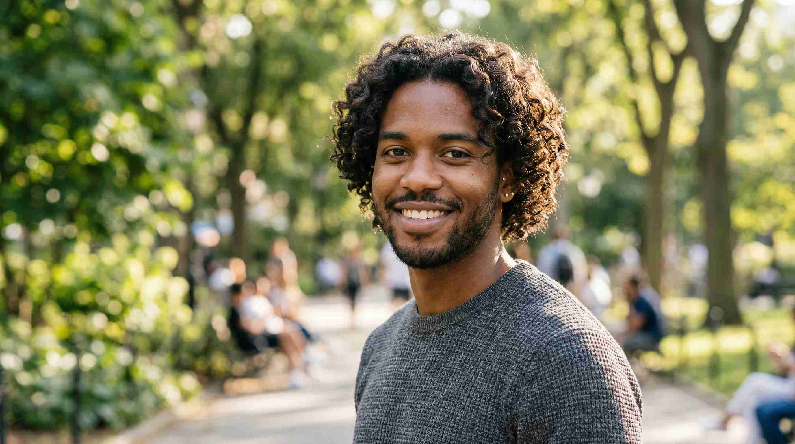 A man with curly hair stands in front of a cafe, casually dressed and looking towards the camera.