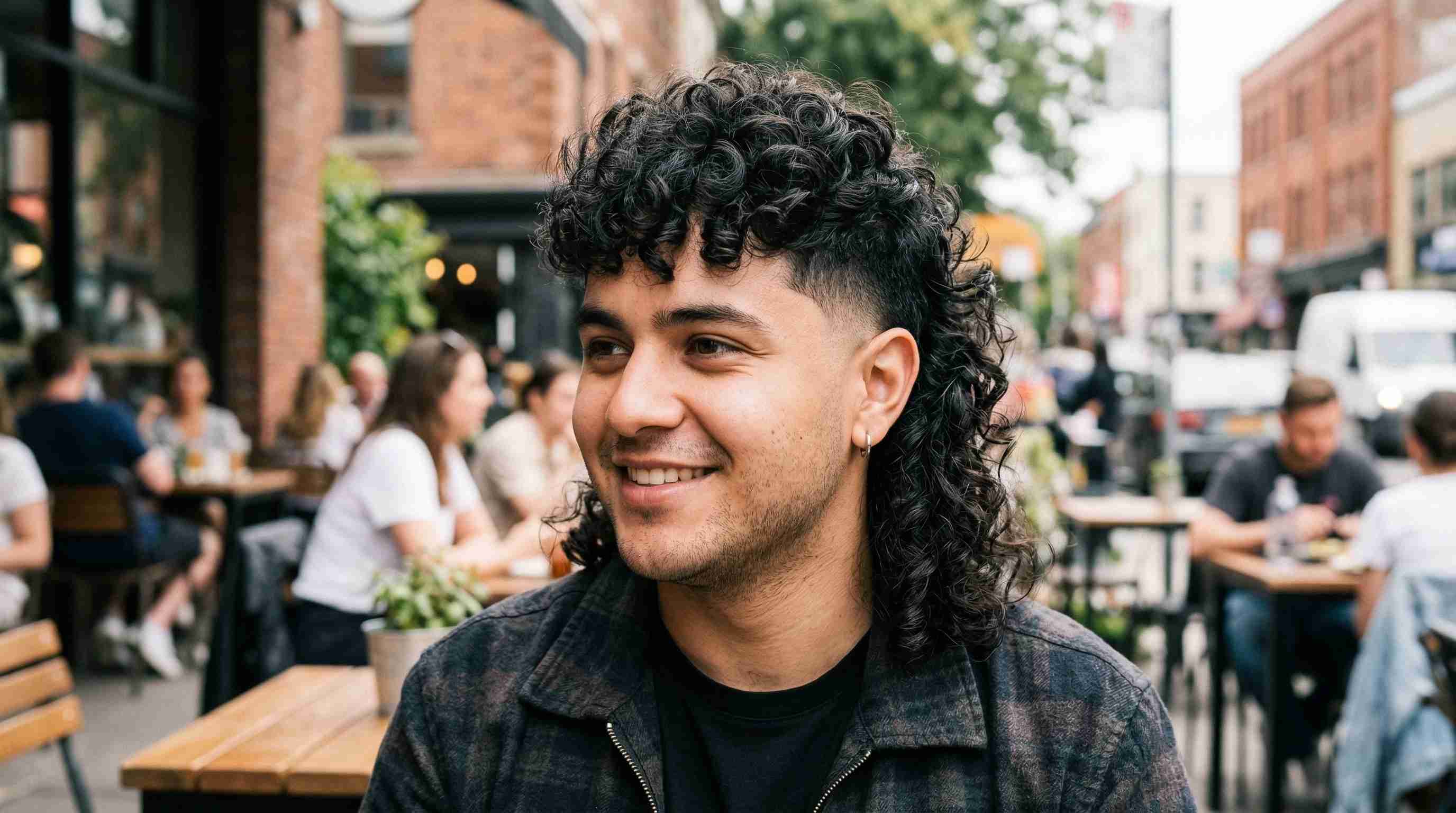 Burst Fade Mullet Round Face UK: 5 Styles & Arc Heights 5 A man with curly hair and a round face sitting at a table, showcasing a burst fade mullet hairstyle.